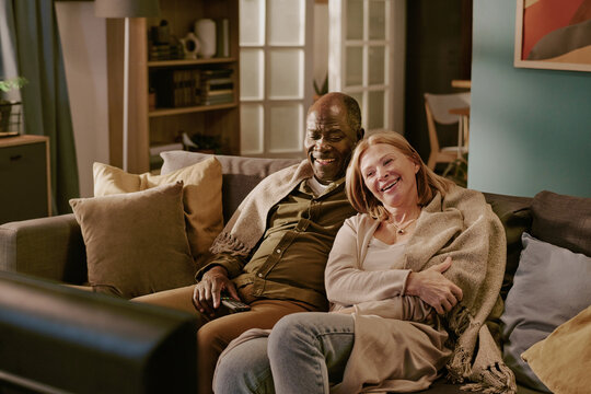 Senior Caucasian woman and senior Black man sitting close together on sofa smiling and watching television, woman leaning affectionately on man, couple sharing romantic moment at home - Powered by Adobe