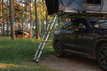 A modern SUV with a rooftop tent and ladder is parked in a forest of tall pine trees. Sunlight filters through, with a wooden cabin in the background.