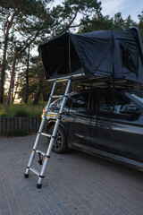 A modern SUV parked on a paved surface in a forest, featuring a black rooftop tent with a telescopic ladder. Tall trees and sunlight create a serene setting.