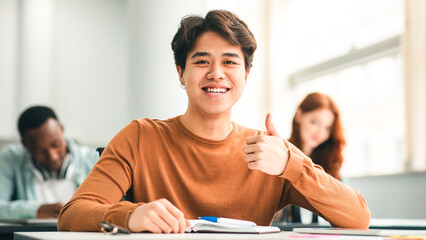 Education Concept. Portrait of smiling asian male student sitting at desk in classroom at university, showing thumbs up sign gesture, looking posing at camera, selective focus. Academic Success © Prostock-studio