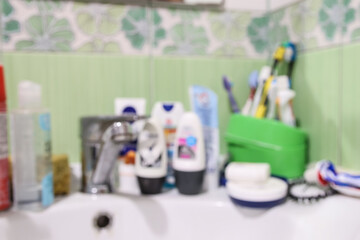 A blurred and messy bathroom sink, featuring an antiperspirant, toothbrush, toothpaste, and soap near the faucet. Bathroom mess on a sink with a blurred background