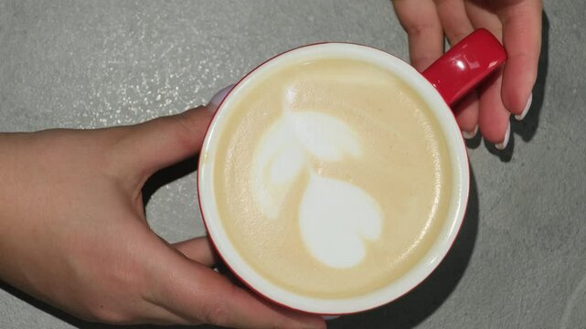 Female barista with nails turns around cup of coffee in hands. Woman puts drink with pattern made of milk in form of flower on table