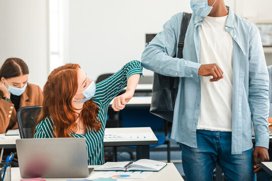 New Normal Greeting. Young multiethnic students wearing protective disposable surgical masks greeting each other at classroom. Black guy standing, redhead woman sitting at desk, people avoid touching