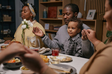 Black family with child holding hands while praying before festive meal enjoying dinner together, sitting around table with delicious food in living room