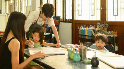 A preschool teacher engages young children in a fun painting session, with a mother also...