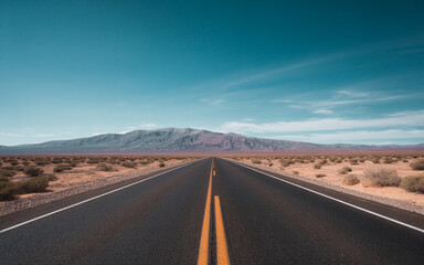 A straight asphalt road in the desert leads toward a distant mountain range under a bright blue sky with soft clouds.