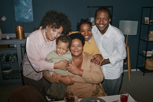 Portrait of happy multigenerational family gently embracing while posing at dinner table during mealtime gathering in living room, camera flash