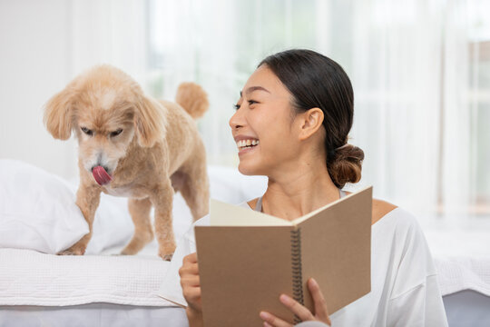Asian Joyful woman reading book with cute dog on bed, smiling happily in cozy bright room, enjoying leisure time together