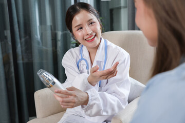Asian smiling female doctor explaining skincare treatment to patient, holding skincare device in...