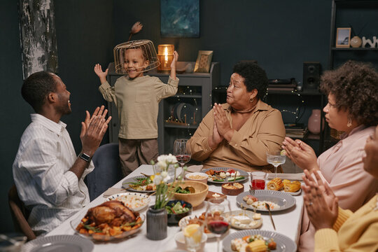 Black family sharing food and laughter around dinner table enjoying festive meal together while child entertaining adults by playing with basket on head - Powered by Adobe