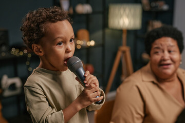 Young Black boy singing into microphone with expressive face while sitting next to elder family...