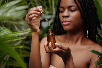 Woman holding dropper bottle of botanical oil serum while surrounded by lush green leaves, focusing...