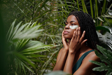 Woman with face mask and eye patches enjoying rejuvenating moment surrounded by lush greenery,...