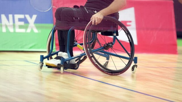 A man in a wheelchair is holding a tennis racket. The court is red and white. There are two banners on the court, one of which says BWF