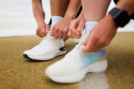 Two Athletes Prepare for a Morning Run by Tying Their Shoelaces on a Sandy Track Near the Beach During a Sunny Day - Powered by Adobe