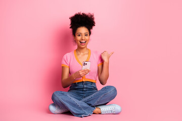 Young woman sitting against pink background holding smartphone smiling and pointing, showcasing...