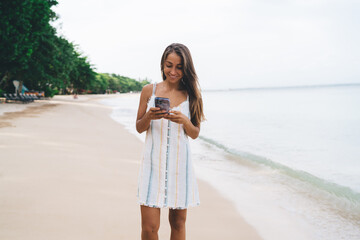 Young Caucasian woman enjoying barefoot stroll along tropical beach holding phone, smiling peacefully while appreciating ocean view and freedom