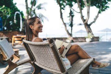 Side profile of joyful woman holding notebook and pen, smiling at serene view from tropical resort...