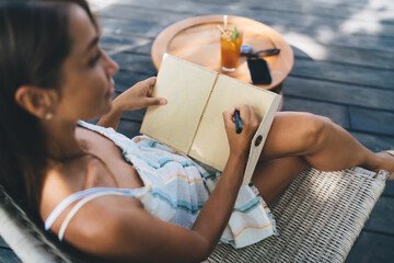 Young female freelancer sketching ideas in blank notebook while seated near phone and sunglasses, prioritizing mindful analog tasks over screen time during work break