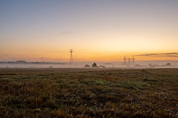 The morning light illuminates a pastoral view where a power transmission tower rises among autumn meadows and fields.