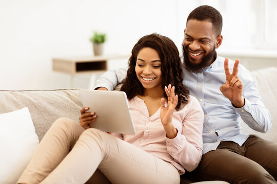 Online Communication. Smiling african american man and woman making digital video chat with friends or family using tablet, waving to webcam, sitting and resting on the couch in living room