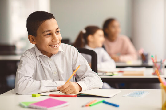 Elementary And Primary School Education. Portrait of smiling small boy sitting at table in classroom, writing or drawing in notebook, looking away at window. Reopening and return back to school