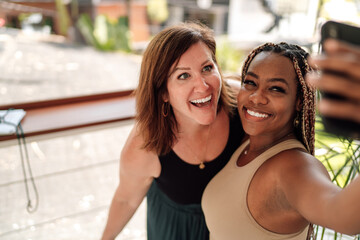 Two happy women pose with phone for selfie, expressing digital closeness, inclusivity and joyful connection in bright urban setting.