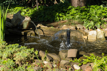 Serene garden pond with small waterfall feature. Water gently cascades from frog statue into pond, surrounded by rocks and lush greenery, creating tranquil and natural atmosphere.