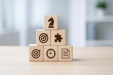 Wooden blocks with business strategy icons including chess knight, target, puzzle, clock, and document stacked on table in bright office setting, symbolizing planning and goal achievement