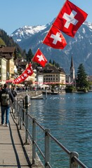 Obraz premium Swiss flags lining a calm lake promenade with distant snow-capped mountains and a picturesque town, representing national pride. Swiss National Day background for greeting card or banner. 