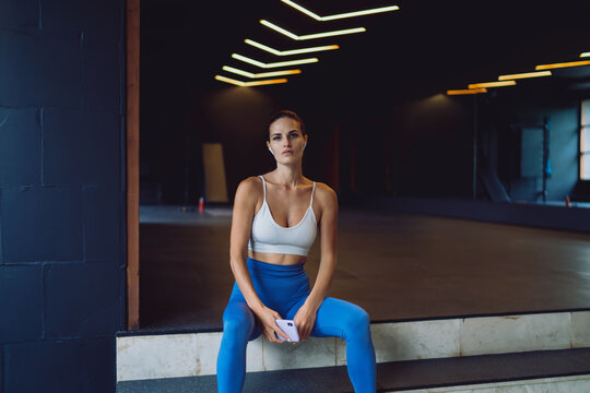 Young woman in gym attire sits on steps holding smartphone, serious expression reflects mental focus, tracking progress, and staying connected through fitness technology and apps.