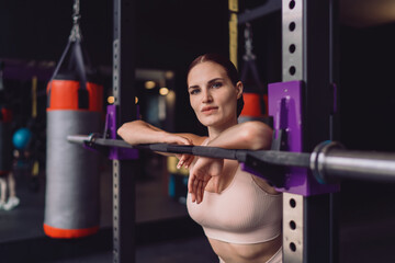 Sporty woman leaning on barbell, direct gaze toward camera, expressing mental toughness,...