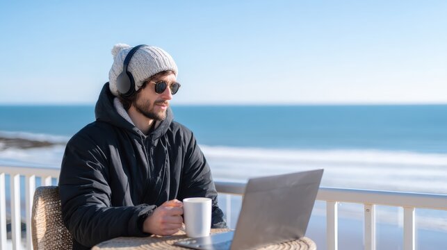A man in winter clothing works on a laptop while holding a coffee mug on a balcony overlooking the ocean on a clear day.