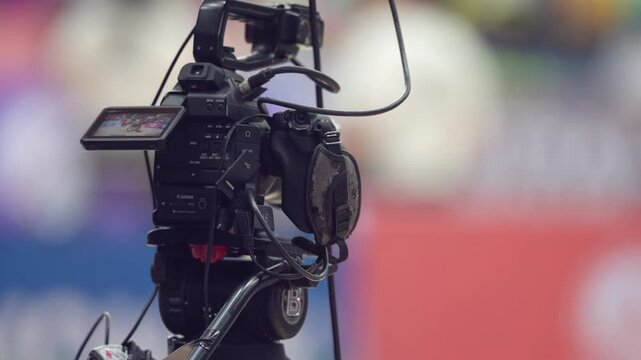 A black camera is mounted on a tripod and is positioned in front of a red and white background. The camera has a red and white screen