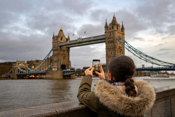 Woman in fur-trimmed coat taking smartphone photo of Tower Bridge during dramatic winter sunset, Thames River view with cloudy sky in London