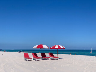 Bright red beach umbrellas and lounge chairs sit on a pristine white sand beach with a vivid blue...