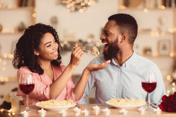 Romantic Relationship. Beautiful lovely young african american couple sitting at table and eating dinner. Smiling woman is feeding her man with pasta. Glasses with red wine and candles on the desk