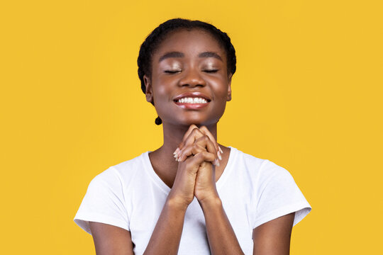 African American Woman Praying Holding Hands In Prayer Gesture Standing On Yellow Studio Background, Smiling With Eyes Closed. Black Female Prays. Hope Concept.