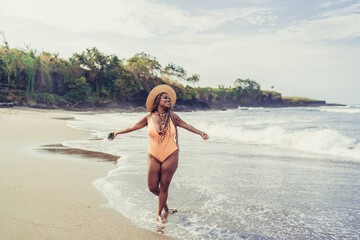 Happy young Black woman stands at ocean’s edge holding phone, radiating empowerment, liberation...