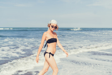 Stylish young woman in black bikini smiling while walking near surf line, portraying authentic joy, confidence and the beauty of spontaneous content moments under natural light.