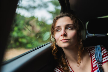 Young digital nomad woman wearing headphones looking out of car window contemplating the landscape