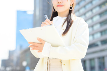 白い服の女性が資料にペンを走らせながら街中で立ち止まり思考する姿
