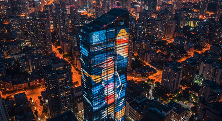An architectural transformation of a skyscraper seen from a high angle, with its facade lit by bold, shifting colors of magenta, blue, and yellow light art.