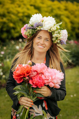 Smiling woman in flower crown holding vibrant peony bouquet, standing in a blooming garden during a...