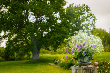A vibrant bouquet of wildflowers in white, purple, and pink set on a stand in a lush garden, with a...