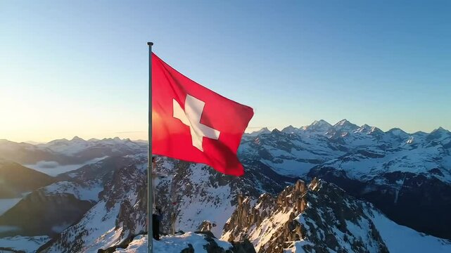 Swiss flag waving atop a snow covered mountain peak with a vast mountain range in the distance