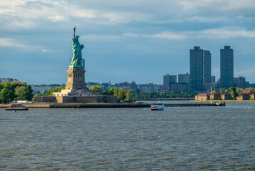 The Statue of Liberty stands tall in New York Harbor, with Jersey City buildings visible in the background.