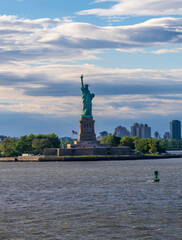 The Statue of Liberty stands tall in New York Harbor, with Jersey City buildings visible in the background.
