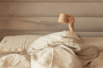 A hand holding a coffee mug emerging from a cozy bed.