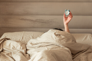 A hand holding an alarm clock emerging from a cozy bed.